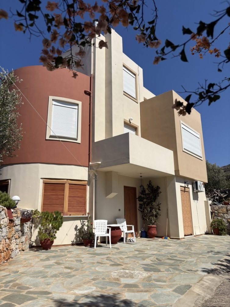 Ancestral Earth guesthouse met aardetinten en houten accenten op Oost-Kreta, omringd door olijfbomen en mediterrane planten.