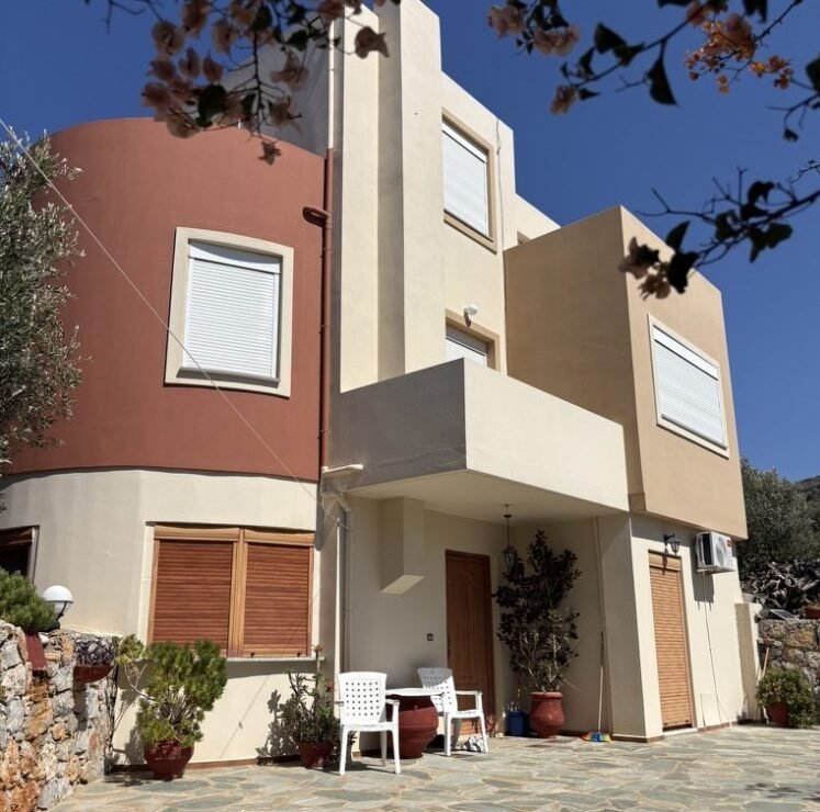 Ancestral Earth guesthouse met aardetinten en houten accenten op Oost-Kreta, omringd door olijfbomen en mediterrane planten.