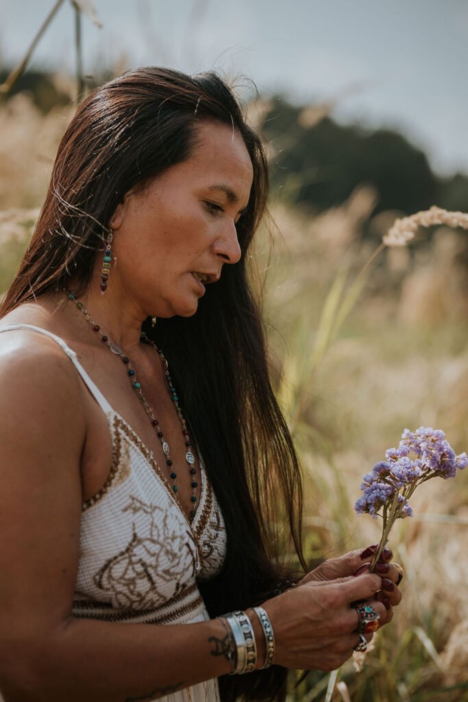 Vrouw in natuurlijke omgeving houdt een paarse bloem vast, symbool voor rust, intuïtie en verbinding met de aarde – sfeerbeeld van Ancestral Earth.